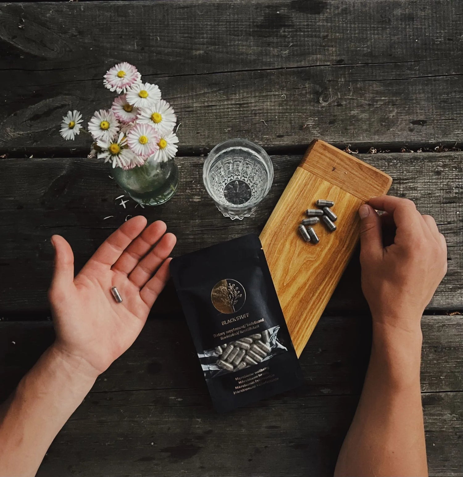 Hands holding and reaching for capsules next to a "BLACK STUFF" dietary supplement pouch, with a glass of water, daisies in a vase, and capsules on a wooden board on a rustic table