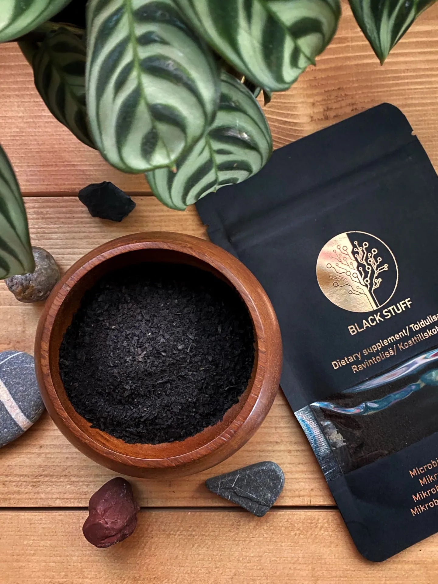Wooden bowl filled with black powder beside a "BLACK STUFF" dietary supplement pouch, surrounded by stones and green leaves on a wooden table