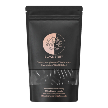 A black resealable pouch labeled "BLACK STUFF" dietary supplement. The packaging features a circular logo with a stylized tree design, and text in multiple languages indicating it is for microbiome wellbeing. The lower part of the pouch is transparent, showing black capsules inside.