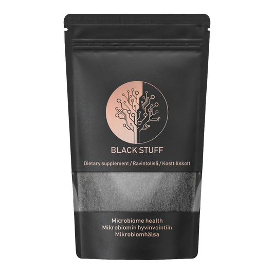A black resealable pouch labeled "BLACK STUFF" with a circular logo resembling a tree made of circuit lines. The packaging states "Dietary supplement" in multiple languages and "Microbiome health" in English, Finnish, and Swedish. The lower part of the pouch has a transparent window showing black powder inside.
